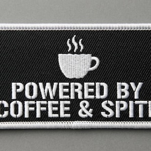 Puede incluir: Un parche rectangular bordado con fondo negro y borde blanco. El diseño presenta una taza de café blanca bordada con vapor elevándose sobre ella, seguido del texto blanco "POWERED BY COFFEE & SPITE" en letras mayúsculas.