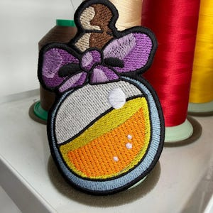 May include: Embroidered patch featuring a potion bottle design. The patch has a gray and orange potion with a purple bow and brown top. The patch is outlined in black and blue. The background includes spools of thread.