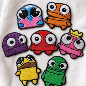May include: A collection of seven embroidered patches featuring cartoon characters in various colors including blue, orange, purple, red, pink with a yellow crown, yellow, and green. Each patch has large white eyes with black pupils.