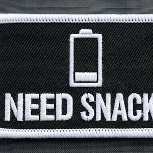 May include: A rectangular embroidered patch with a black background and a white border. The design features a white outline of a low battery icon centered above the white text that reads NEED SNACKS in a bold, sans-serif capital letter font.