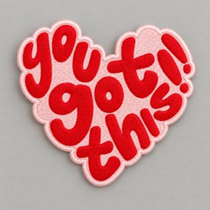 May include: A heart-shaped embroidered patch with a light pink background and bold red lettering that reads "you got this!!" in a rounded, playful font. The edges of the patch are scalloped and stitched in a matching light pink thread.