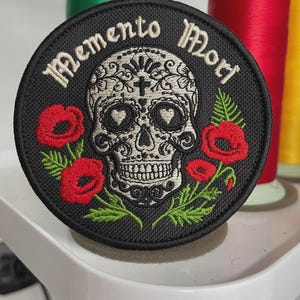 May include: A round black patch embroidered with a sugar skull design, red poppies, and the words "Memento Mori" in white. The skull has heart-shaped eye sockets and intricate floral patterns. Green and red thread spools are in the background.