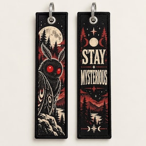 Puede incluir: Dos etiquetas rectangulares de tela negra con anillas de metal plateado. Una etiqueta muestra una polilla negra con ojos rojos sobre un fondo de bosque y luna. La otra muestra el texto STAY MYSTERIOUS con fases lunares y un paisaje forestal.