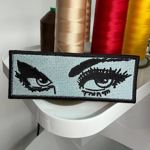 May include: A rectangular patch featuring a stylized black eye design on a light blue background. The patch is bordered in black. Spools of thread in various colors, including beige, red, and yellow, are visible in the background.