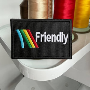 May include: Black rectangular patch with the word "Friendly" in white. The patch features a colorful design with blue, green, yellow, and red curved lines. Spools of thread in various colors are in the background.