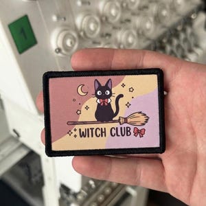 Op de afbeelding: Een rechthoekige geborduurde patch met een zwarte rand, voorzien van een zwarte kat met een rode vlinderdas, zittend op een bezemsteel. De achtergrond heeft golvende vlakken in roze, geel en paars met een maansikkel, sterren en de tekst WITCH CLUB.