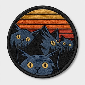 May include: A circular embroidered patch featuring three dark blue cat heads with yellow eyes peeking out from behind black mountain silhouettes. The background displays a horizontal striped sunset pattern in shades of orange and yellow.