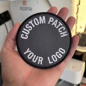 Op de afbeelding: Een ronde geborduurde patch met een donkergrijs midden en een zwarte rand. De tekst "CUSTOM PATCH" is met wit garen langs de bovenste boog geborduurd en de tekst "YOUR LOGO" is met wit garen langs de onderste boog geborduurd.
