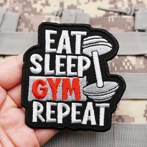 Può includere: Una toppa ricamata nera con il testo bianco EAT SLEEP e REPEAT e il testo rosso GYM al centro. A destra del testo si trova un'icona di un manubrio ricamata in bianco. La toppa presenta un bordo nero e una forma sagomata.