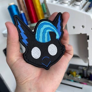 May include: An embroidered patch featuring a stylized black creature head with large white circular eyes, a blue lightning bolt design on the ears, a blue striped crest on the forehead, and a small blue V-shaped mouth stitched on a black background.