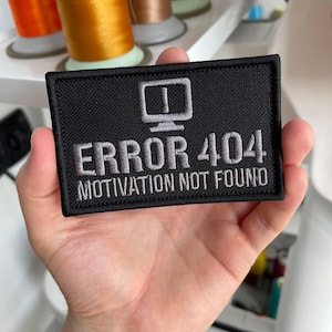 Puede incluir: Un parche rectangular bordado en negro con un icono de monitor de ordenador gris en la parte superior. Debajo del icono, el texto dice ERROR 404 en letras grises grandes, seguido del texto MOTIVATION NOT FOUND en letras grises más pequeñas.