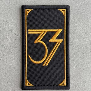 May include: Black rectangular patch with gold embroidered number 33. The number is stylized with a curved bottom and angled lines. The patch has a gold border.