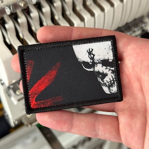 Puede incluir: Un parche rectangular bordado con borde negro. El diseño presenta un fondo negro a la izquierda con una marca de pincelada roja y un gráfico de calavera blanca a la derecha con un símbolo negro en la frente.