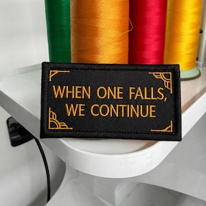 以下が含まれることがあります： 「WHEN ONE FALLS, WE CONTINUE」という金色の文字と金色の縁取りが施された、長方形の黒い刺繍ワッペン。ワッペンの四隅には金色の装飾的なアクセントがあり、カラフルな糸巻きの前に配置されています。