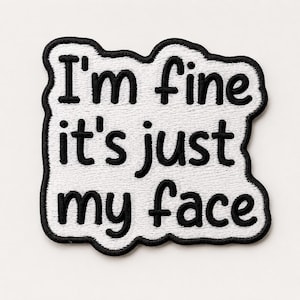 Puede incluir: Un parche bordado blanco con un borde negro y el texto negro "I'm fine it's just my face" en una fuente redondeada y negrita. El parche tiene una forma irregular y orgánica que sigue el contorno de las letras, creando un diseño de alto contraste.
