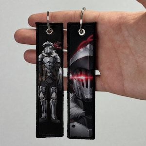Puede incluir: Dos llaveros rectangulares de tela con fondos negros e ilustraciones estilo anime de un guerrero con armadura plateada. Uno muestra al personaje de cuerpo entero, mientras que el otro presenta un primer plano de un casco con ojos rojos brillantes.