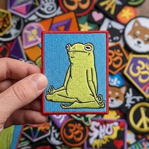 May include: A rectangular embroidered patch featuring a light green frog sitting in a cross-legged meditation pose with hands in mudra. The frog is set against a solid light blue background and is framed by a bold, stitched red border.
