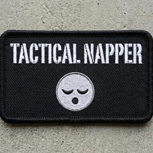 May include: A rectangular black embroidered patch with a stitched border. The text TACTICAL NAPPER is embroidered in white capital letters at the top. Below the text is a circular white face icon with closed eyes and an open mouth in black thread.