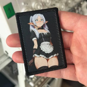 May include: A rectangular black patch featuring a cartoon illustration of a female anime character in a maid outfit. The character has white hair, pointed ears, and is wearing a black dress with a white apron. The patch has a stitched border.