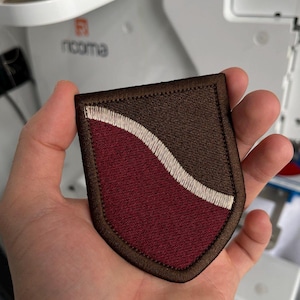 Op de afbeelding: Een bruin en bordeauxrood geborduurd embleem met een wit golfontwerp. De patch heeft een schildvorm met een bruine rand en wordt in een hand gehouden. De patch is waarschijnlijk voor kleding of accessoires.