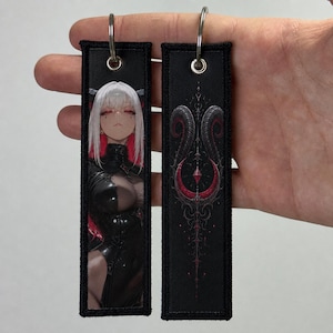 May include: Two rectangular embroidered fabric keychains with silver metal rings. One keychain features an anime-style character with white hair and red accents in a black outfit. The other displays a symmetrical black and red horned emblem.