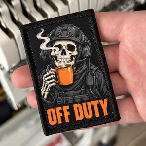Könnte beinhalten: Ein rechteckiger, bestickter Aufnäher mit schwarzem Rand, der ein Skelett mit taktischem Helm und Headset zeigt, das eine orangefarbene Kaffeetasse mit aufsteigendem Dampf hält. Der Text OFF DUTY ist unten in Orange aufgedruckt.