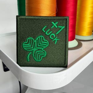 Puede incluir: Un parche cuadrado bordado en color verde oliva con un diseño de trébol verde brillante y el texto +1 LUCK cosido en hilo verde a juego. El parche tiene un borde grueso cosido y se muestra sobre un fondo de carretes de hilo de colores.