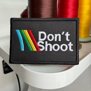 May include: A black rectangular patch with the text "Don't Shoot" in white. The patch features a colorful design of blue, yellow, green, and red stripes. The patch is likely an embroidered item.
