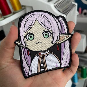 May include: Embroidered patch depicting an anime-style girl with light purple pigtails, green eyes, and pointed ears. The patch has a black border and is held in a hand. The girl is wearing a white and black outfit.