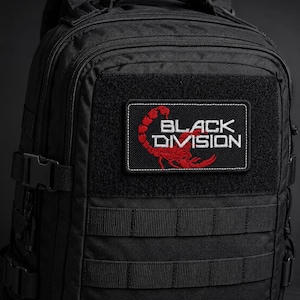 May include: A black tactical backpack featuring a rectangular patch on the front. The patch displays a red scorpion graphic and the white text "BLACK DIVISION" embroidered on a black background, with multiple horizontal webbing straps below.