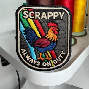 Op de afbeelding: Geborduurde patch met een haanontwerp. De patch heeft de tekst "SCRAPPY ALWAYS ON DUTY" in witte letters op een zwarte achtergrond. De haan is afgebeeld in levendige kleuren, waaronder rood, geel, blauw en groen.