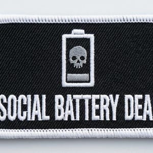Può includere: Una toppa rettangolare ricamata con sfondo nero e bordo bianco. Il design presenta un'icona di batteria bianca contenente un teschio grigio, con il testo SOCIAL BATTERY DEAD ricamato in lettere maiuscole bianche sotto l'icona.