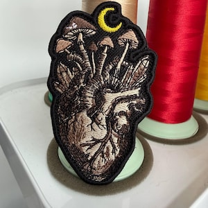 Puede incluir: Parche bordado con un diseño de corazón anatómico, setas y una luna creciente amarilla. El parche está en tonos marrones y negros, con costuras intrincadas. Un accesorio único para chaquetas, bolsos u otros artículos de tela.