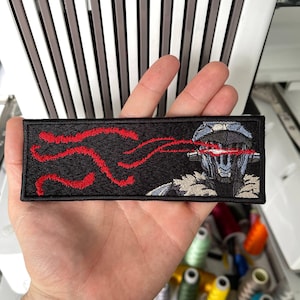 以下が含まれることがあります： 赤いレーザー光線を持つロボットの姿が刺繍された黒い長方形のパッチ。ロボットはグレーとブルーのデザインです。パッチは手で持たれており、背景には糸巻きがあります。