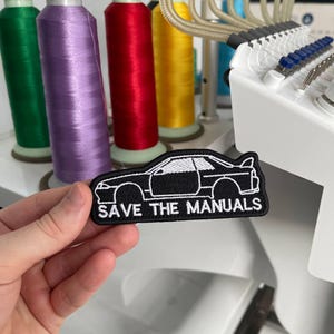 May include: A black embroidered patch featuring a white outline of a sports car silhouette and the text "SAVE THE MANUALS" in white capital letters. The patch is rectangular with rounded edges and is held against a background of colourful thread spools.