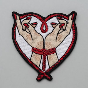 Può includere: Toppa ricamata a forma di cuore, raffigurante due mani con unghie rosse, legate con una corda rossa. Il cuore è bordato di nero con un bordo rosso, su sfondo bianco.