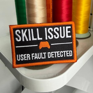 Puede incluir: Un parche rectangular bordado con fondo negro y borde naranja brillante. El texto dice SKILL ISSUE en blanco, seguido de un pequeño icono de mando de juego naranja y el texto USER FAULT DETECTED en letras blancas.