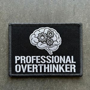 Pode incluir: Um emblema retangular bordado em preto com o contorno branco de um cérebro humano com três pequenas engrenagens no interior. Abaixo do cérebro, o texto PROFESSIONAL OVERTHINKER está bordado em letras maiúsculas brancas com uma borda preta.
