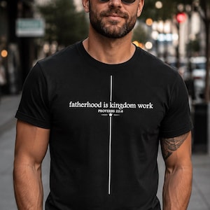 Op de afbeelding: Zwart T-shirt met ronde hals met de tekst "fatherhood is kingdom work" en een verticaal wit lijndesign. De tekst bevat het bijbelvers Spreuken 22:6. Het T-shirt wordt gedragen door een persoon met een zonnebril.