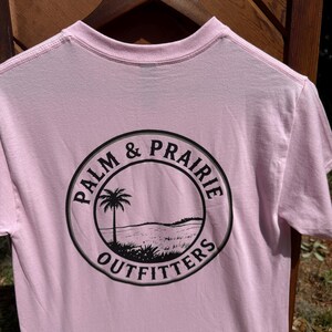 May include: A light pink t-shirt with a crew neck and short sleeves. The back of the shirt features a black circular graphic with the words "PALM & PRAIRIE OUTFITTERS" surrounding a landscape illustration of a palm tree and a field.