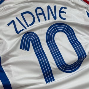 May include: White soccer jersey with blue accents, featuring the name "ZIDANE" and the number "10" in blue. The jersey has red and blue trim on the collar and shoulders. The fabric has a subtle sheen.