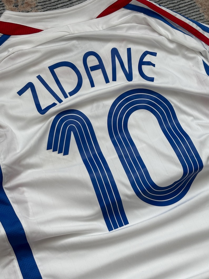 May include: White soccer jersey with blue accents, featuring the name "ZIDANE" and the number "10" in blue. The jersey has red and blue trim on the collar and shoulders. The fabric has a subtle sheen.