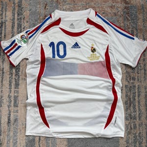 May include: White soccer jersey with red and blue accents. Features the number 10 in blue, the Adidas logo, and a French emblem. Includes stripes on the sleeves and a small patch. The jersey is made of a lightweight, breathable material.