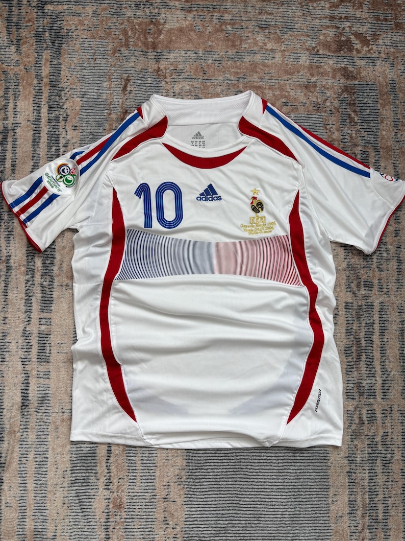 May include: White soccer jersey with red and blue accents. Features the number 10 in blue, the Adidas logo, and a French emblem. Includes stripes on the sleeves and a small patch. The jersey is made of a lightweight, breathable material.