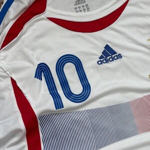 May include: White Adidas soccer jersey with blue and red accents. Features the number 10 in blue on the front, along with the Adidas logo. The jersey has a crew neck and is made of a breathable fabric.
