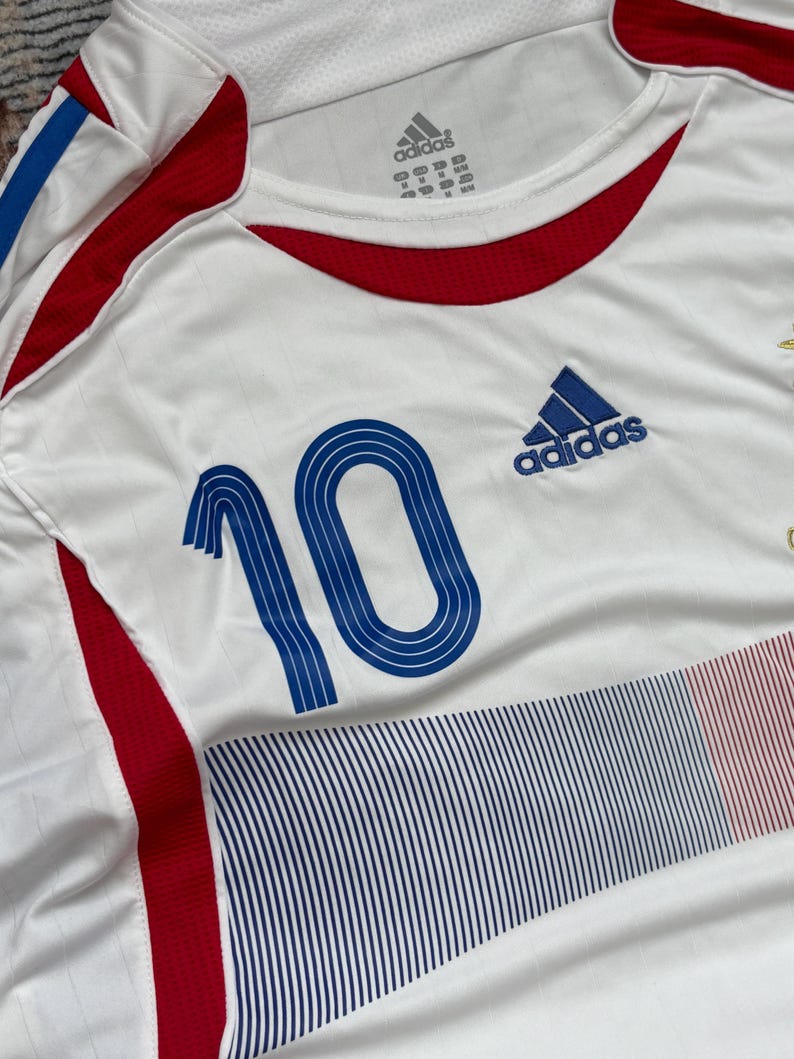 May include: White Adidas soccer jersey with blue and red accents. Features the number 10 in blue on the front, along with the Adidas logo. The jersey has a crew neck and is made of a breathable fabric.