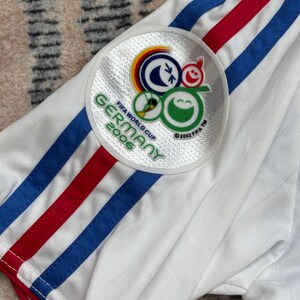 May include: White soccer jersey with blue and red stripes on the sleeve. A circular patch on the sleeve features the FIFA World Cup Germany 2006 logo. The jersey has a ribbed texture and red trim.