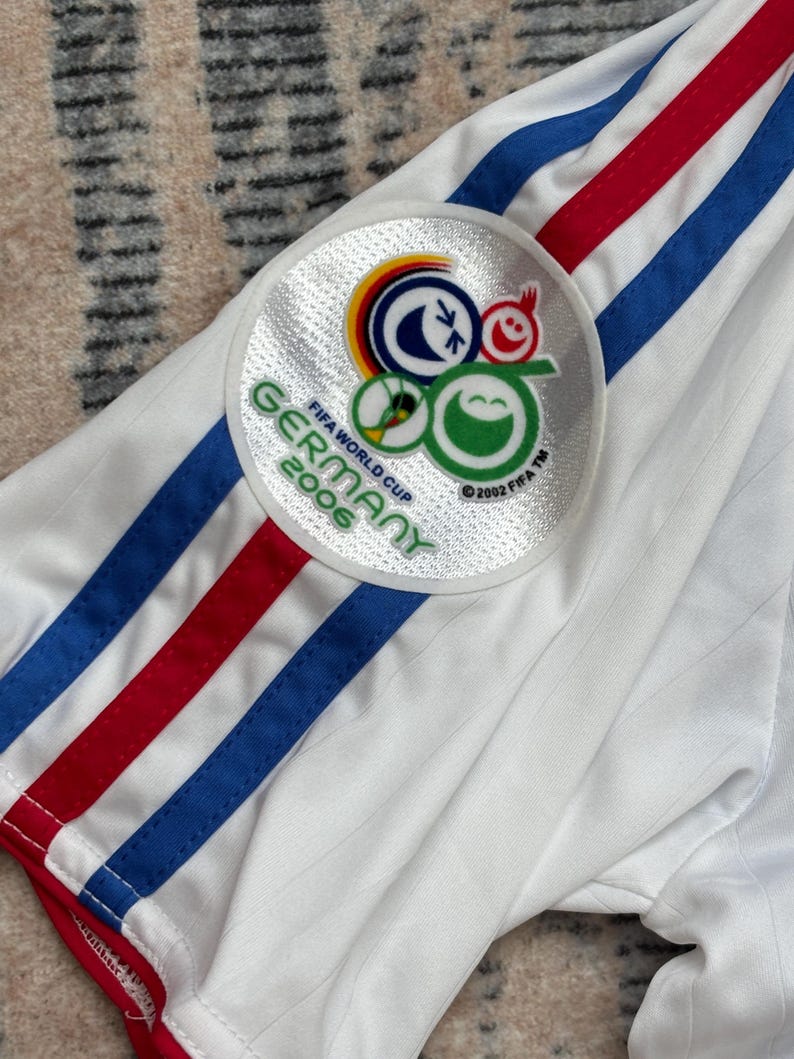 May include: White soccer jersey with blue and red stripes on the sleeve. A circular patch on the sleeve features the FIFA World Cup Germany 2006 logo. The jersey has a ribbed texture and red trim.