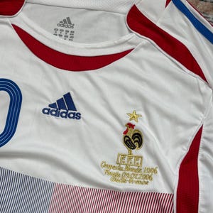 May include: White Adidas soccer jersey with blue and red accents, featuring the French Football Federation logo and the text "Coupe du Monde 2006 Finale 09.07.2006 Italie-France". The jersey has the Adidas logo and a blue number 10.