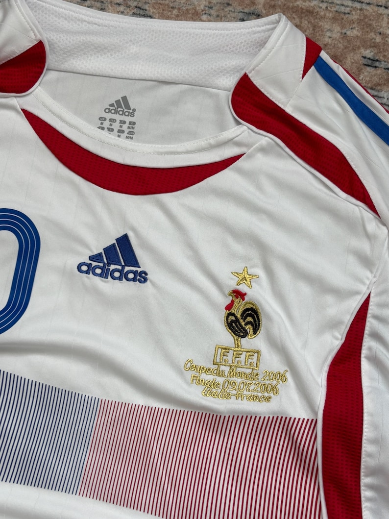 May include: White Adidas soccer jersey with blue and red accents, featuring the French Football Federation logo and the text "Coupe du Monde 2006 Finale 09.07.2006 Italie-France". The jersey has the Adidas logo and a blue number 10.